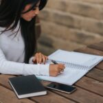 crop ethnic freelancer writing in notebook at street table