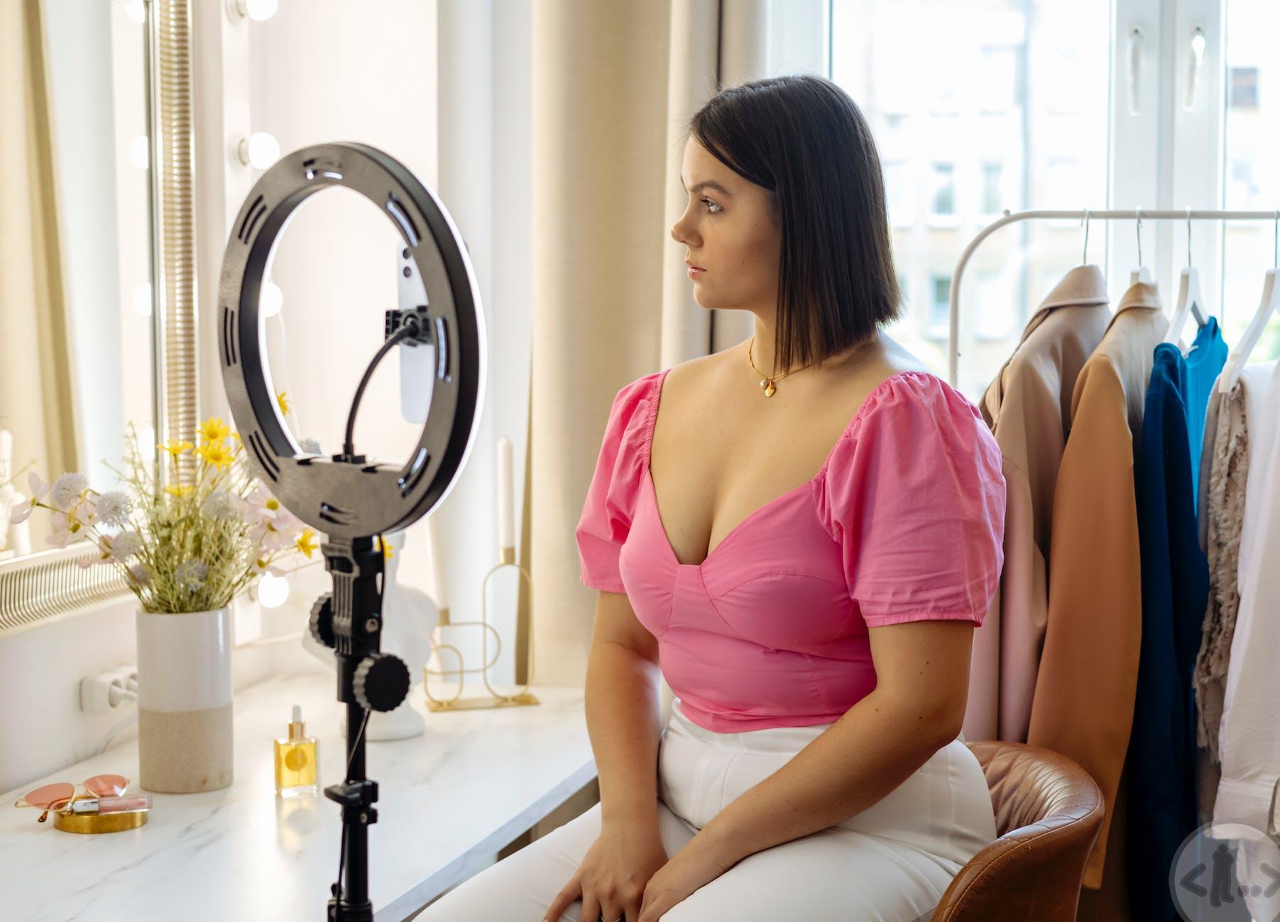 a woman in pink top sitting on brown chair in front of a ring light